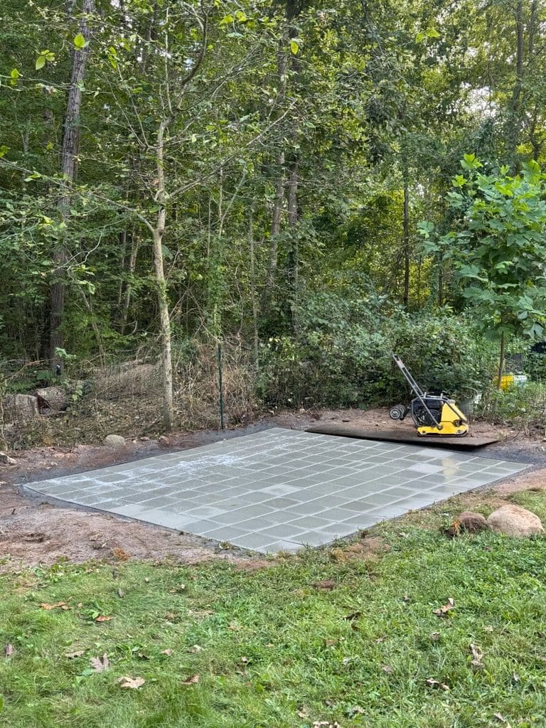 Paved garden area in a wooded backyard, with a compactor ready for landscaping work.