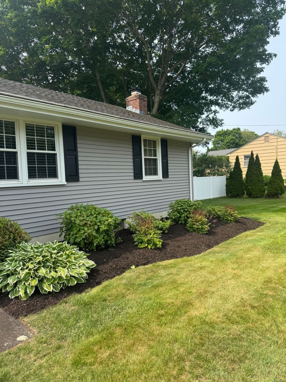Side view of a gray house with black shutters, garden beds, and well-manicured lawn.