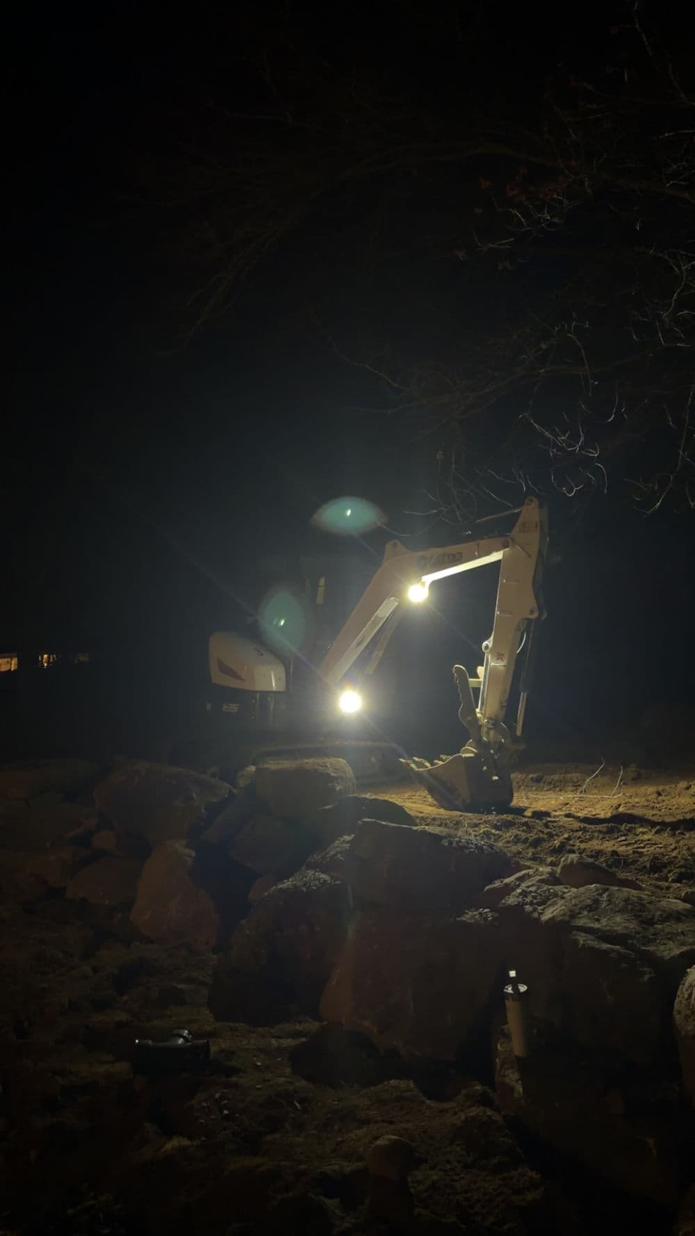 Excavator working at night, illuminated by headlights, with rocks in the foreground.