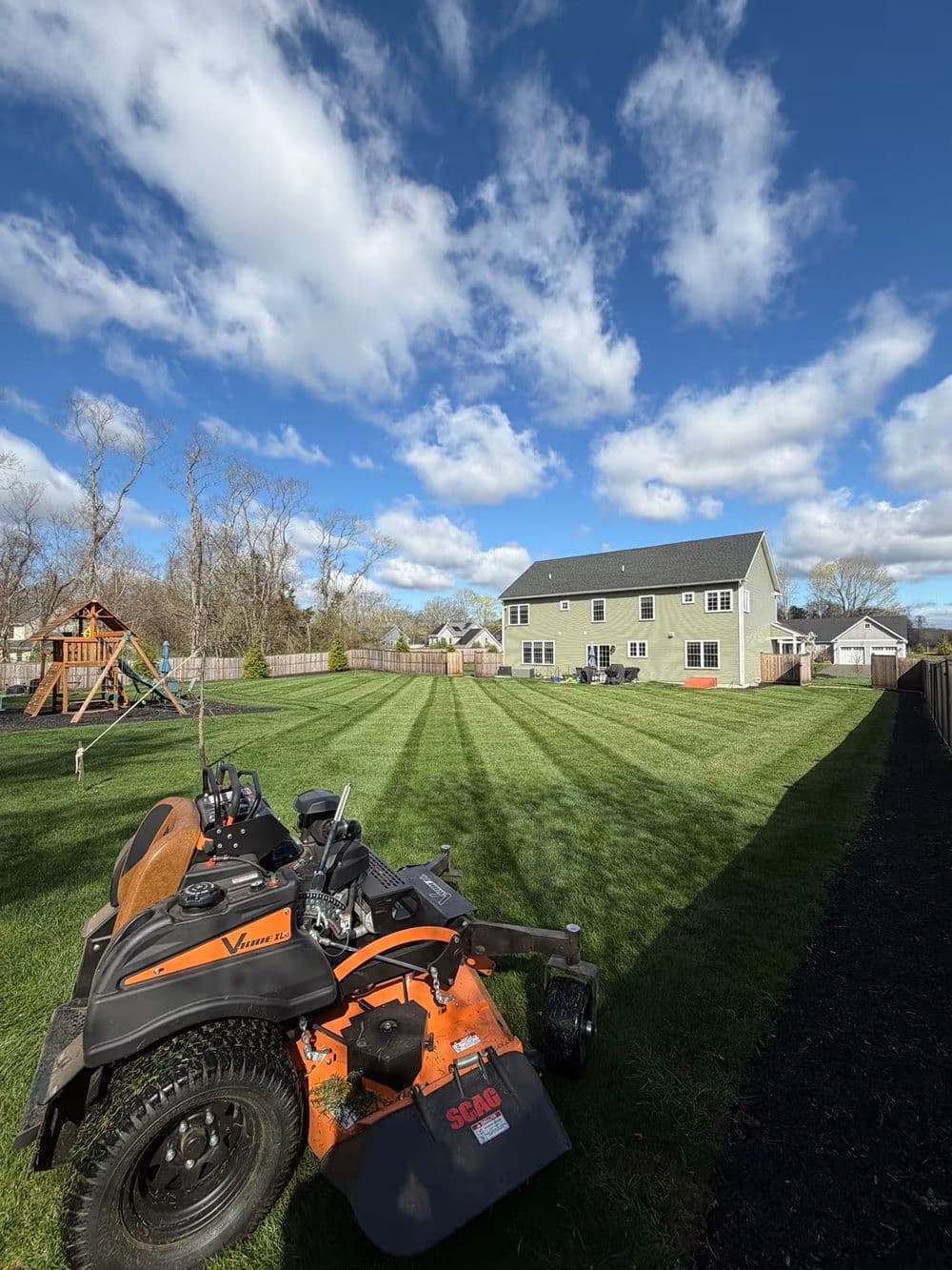 Lawn mower on freshly mowed lawn with playset and house under a blue sky.