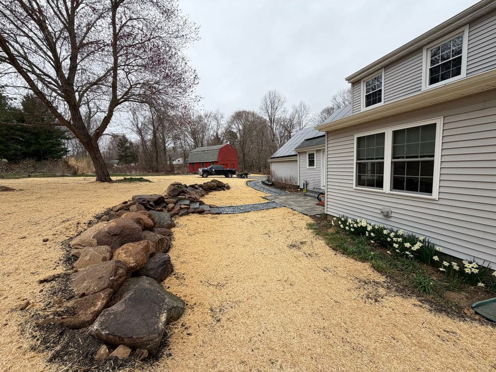 Side yard landscape with stones, pathway, and barn in the background on a cloudy day.