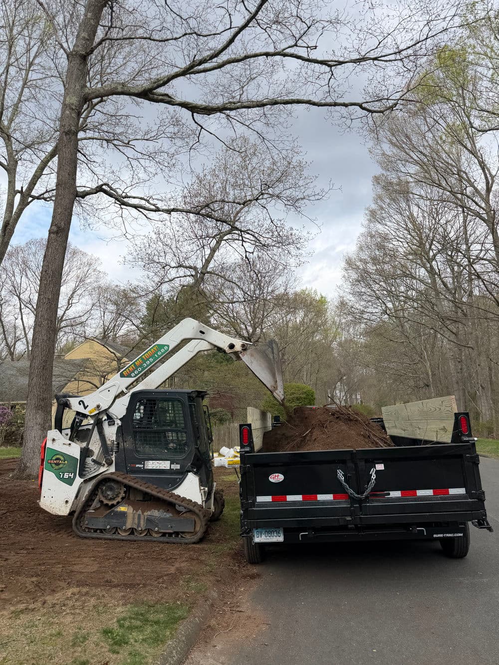 Skid steer loader unloading soil from a trailer in a residential area.